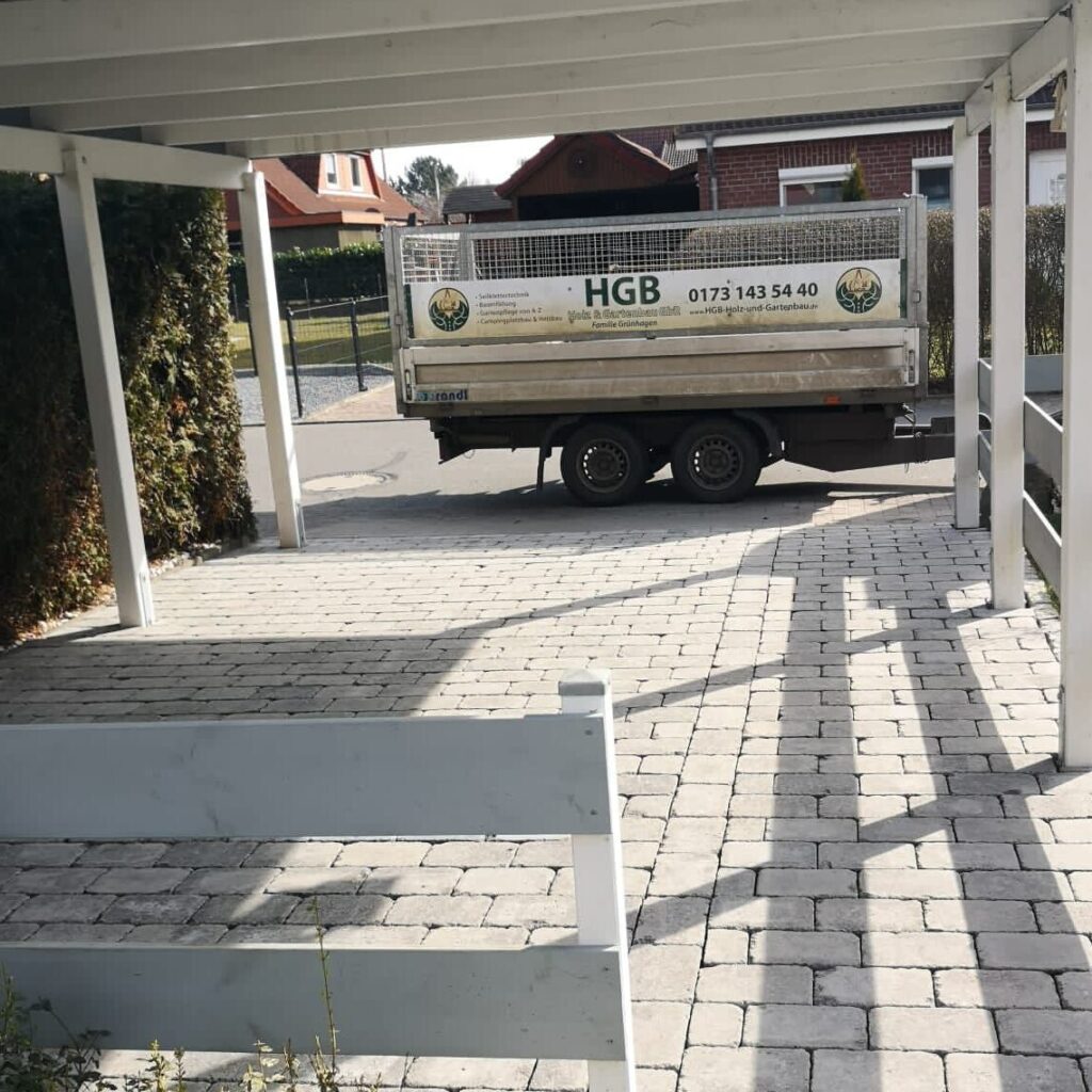Ein Anhänger der HGB Holz und Gartenbau GbR mit Firmenlogo und Kontaktdaten steht auf einer Straße, gesehen von einem gepflasterten Bereich unter einem Carport.