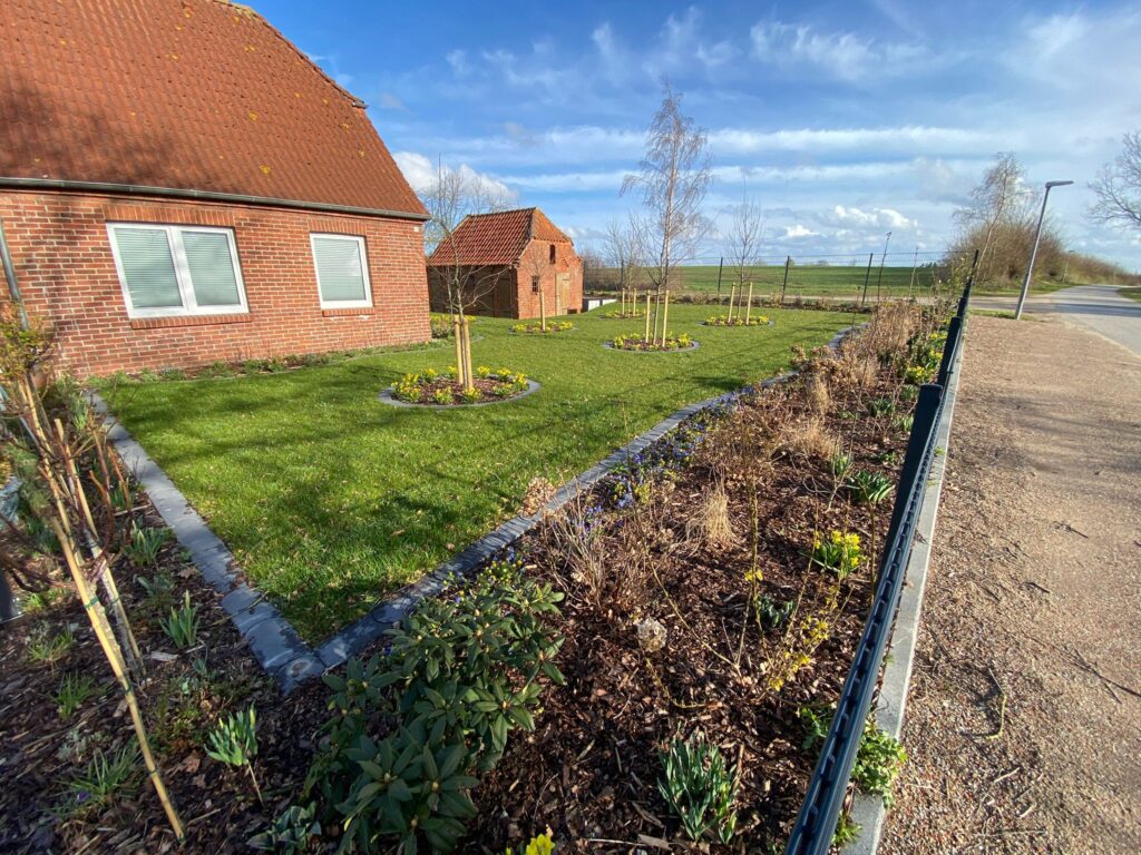 Gepflegte Gartenanlage mit Rasenfläche, jungen Bäumen und Blumenbeeten vor einem roten Backsteinhaus.