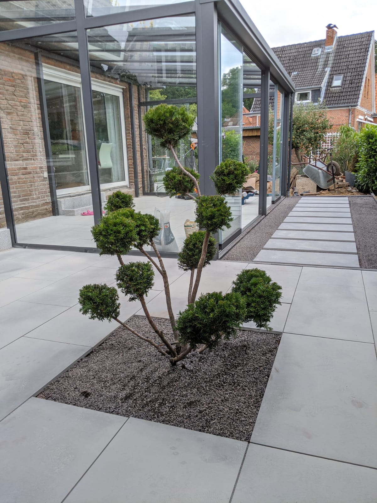 Ein modern gestalteter Terrassenbereich mit großformatigen hellgrauen Betonplatten. In der Mitte wächst ein formgeschnittener Strauch in einem quadratischen Pflanzfeld mit dunklem Kies
