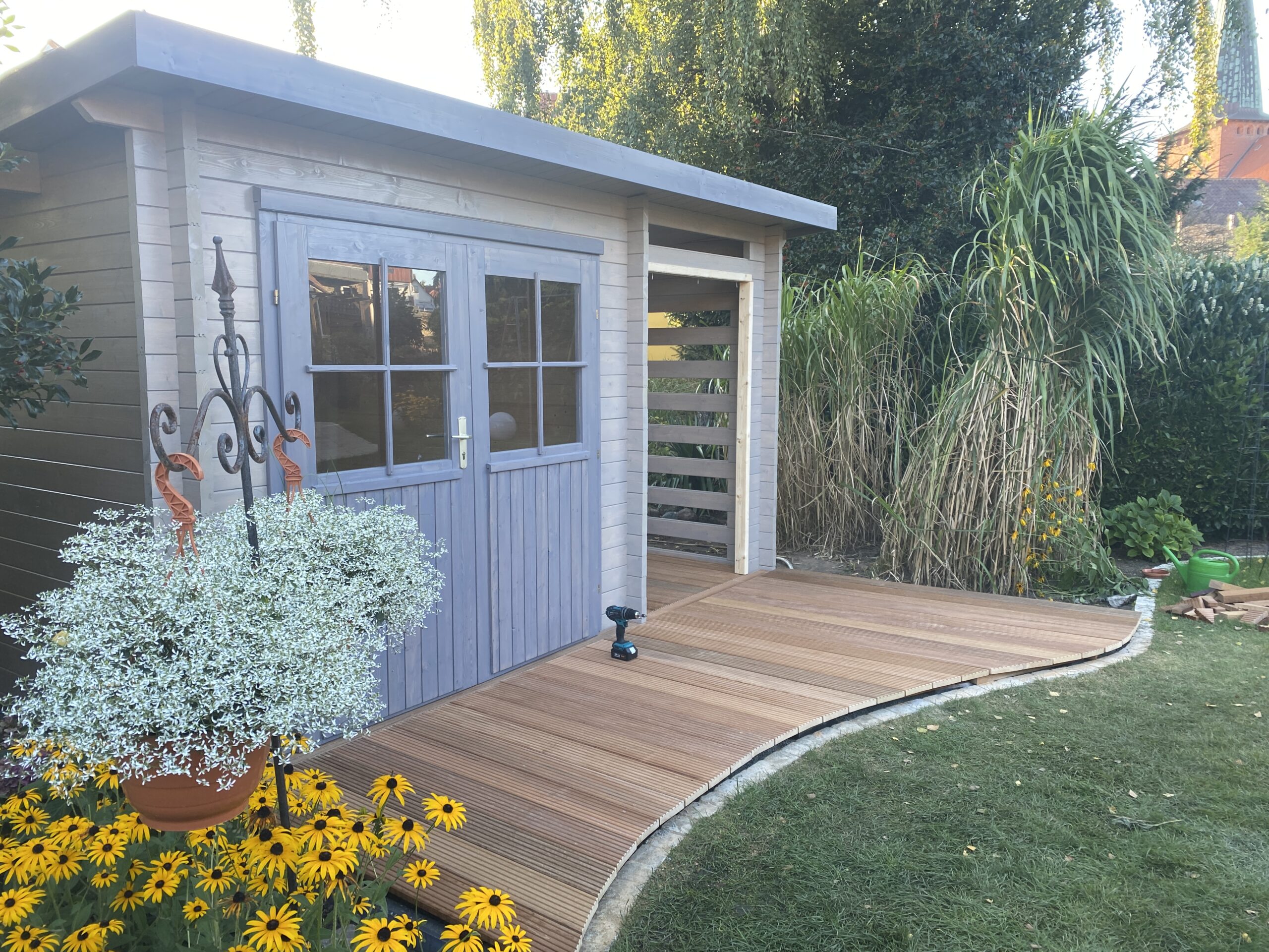 Gartenhaus aus graublau lasiertem Holz mit Doppeltür und Fensterverglasung, davor eine gebogene Terrasse aus Holzbohlen.