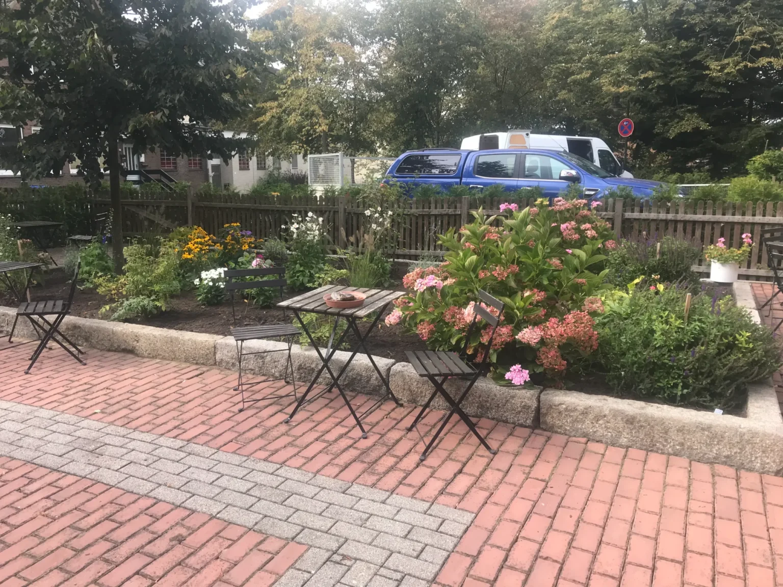 Eine blühende Beetanlage mit Hortensien und Rudbeckien neben einem gepflasterten Terrassenbereich mit Bistrotischen und Stühlen vor dem Hotel Lindenhof.