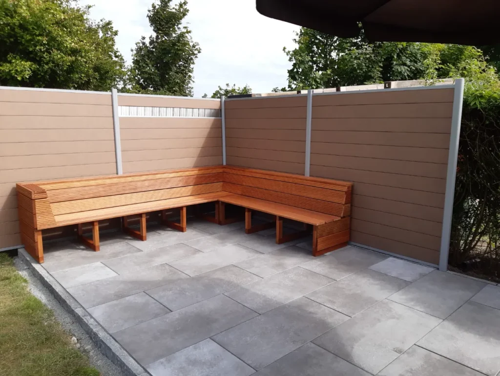 L-förmige Holzbank auf einer gepflasterten Terrasse, umgeben von einem braunen WPC-Zaun und grünen Pflanzen im Hintergrund.
