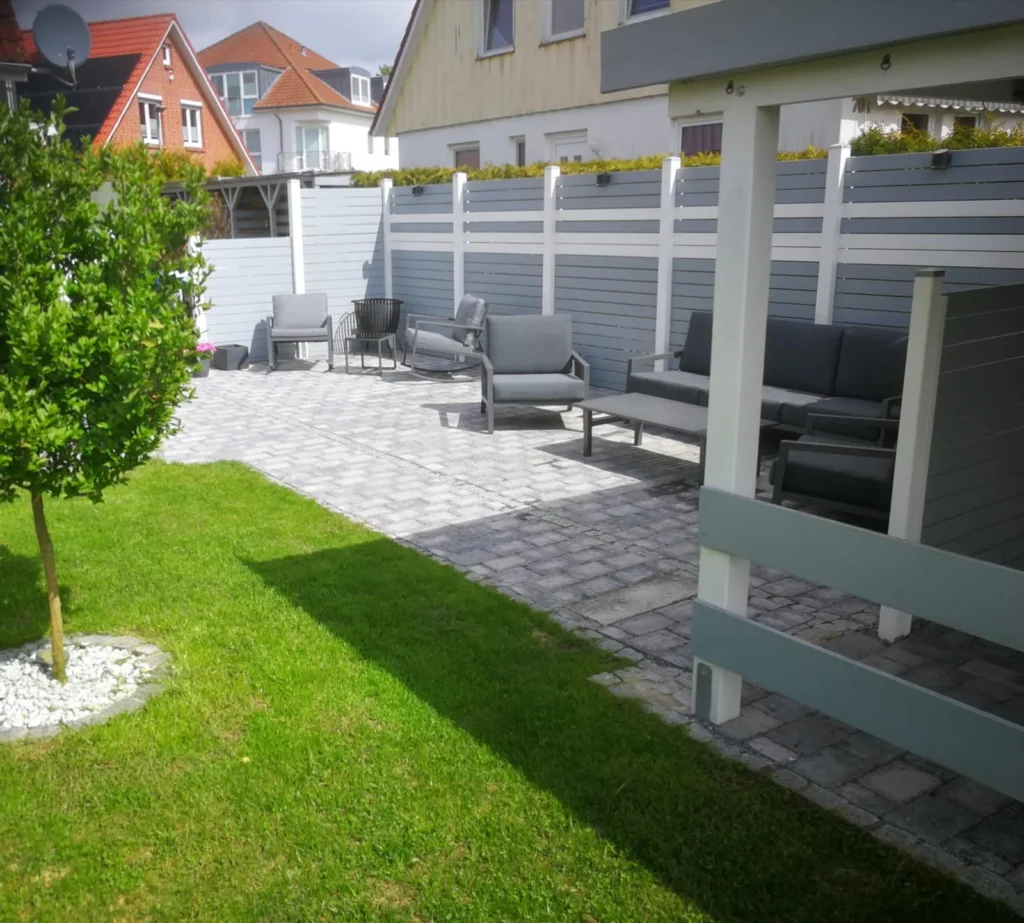 Ein modern gestalteter Garten mit gepflasterter Terrasse, grauen Outdoor-Möbeln, einem Sichtschutzzaun und einer grünen Rasenfläche mit kleinem Baum.