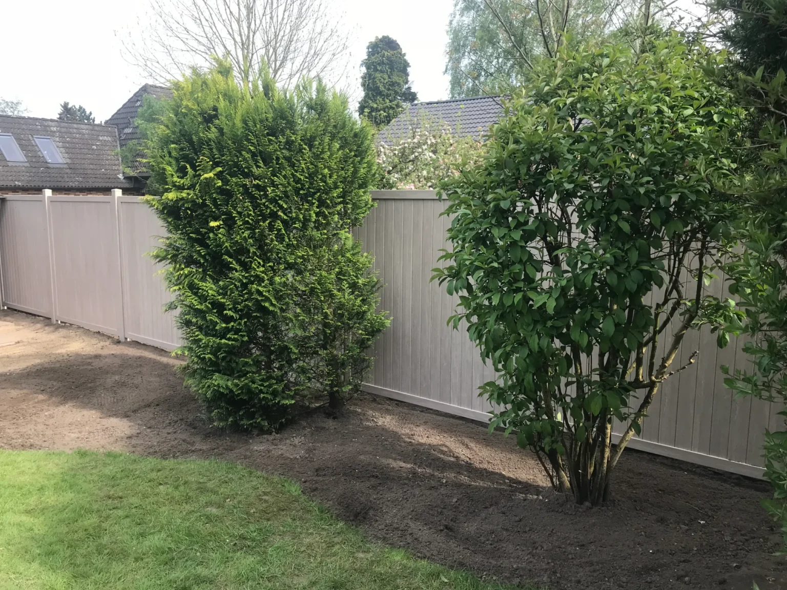 Ein moderner, grauer WPC-Zaun trennt den Gartenbereich mit frisch bearbeiteter Erde und grünen Büschen von einem Wohnhaus im Hintergrund.