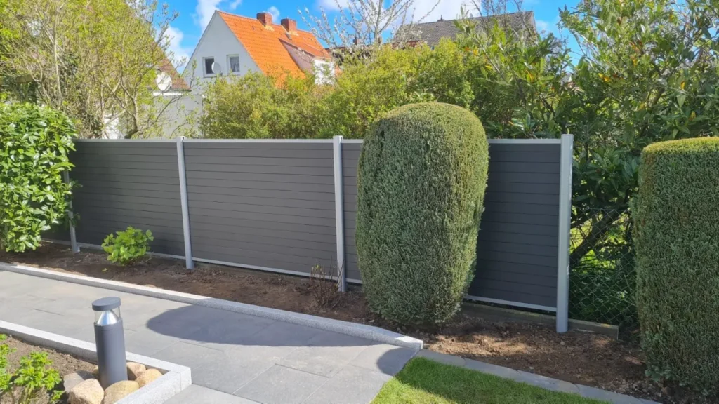 Moderner Sichtschutzzaun aus dunklem Verbundmaterial mit hellen Pfosten in einem gepflegten Gartenbereich mit gepflastertem Weg und grünen Pflanzen.